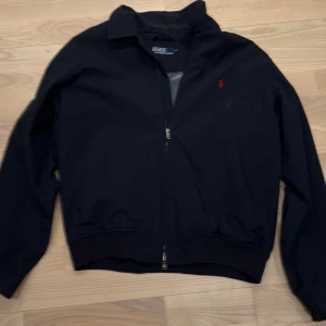 Svart vindjacka från Polo Ralph Lauren - Mörkblå vindjacka från Polo Ralph Lauren med röd broderad logga på bröstet. Jackan har dragkedja framtill, klassisk krage och två sidofickor. Insidan har ett rutigt meshfoder för extra komfort. Perfekt för dig som gillar stilren och sportig look.