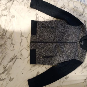 Grå cardigan Abercrombie & Fitch  - Snygg gråmelerad cardigan från Abercrombie & Fitch i storlek S. Jackan har svarta ärmar, ribbade muddar och två dragkedjefickor framtill. Insidan är quiltad för extra komfort och värme. Perfekt för dig som gillar stilren och urban look.