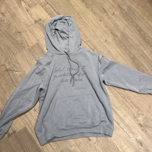 Grå hoodie från H&M med text - Snygg grå hoodie från H&M i storlek L med broderad text på bröstet. Tröjan har huva med snörning och en stor magficka framtill. Perfekt för chill dagar och har en skön, mjuk insida i bomullsmix.