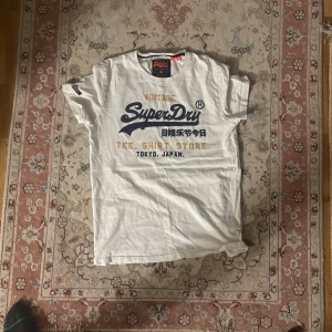 Vit Superdry t-shirt med tryck - Vit t-shirt från Superdry med stort vintage-tryck i marinblått och guld på bröstet. Klassisk rund hals och korta ärmar. Mjuk bomullskvalitet och snygga detaljer som etikett på ärmen. Perfekt för dig som gillar streetwear och japansk-inspirerad design.