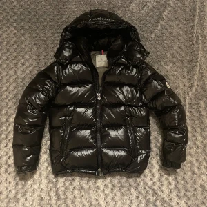 Moncler Maya - Säljer en svart, glansig pufferjacka från Moncler med huva och dragkedja framtill. Jackan har Moncler-logga på ärmen, praktiska fickor med dragkedja och ett coolt serietryck på insidan. Perfekt för dig som vill ha en varm och snygg jacka med streetkänsla.