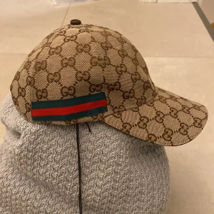 Gucci keps med GG-mönster och ränder - Snygg keps från Gucci i beige och brunt med klassiskt GG-monogram över hela kepsen. På sidorna finns gröna och röda ränder som ger en ikonisk touch. Justerbar rem bak i brunt skinn. Perfekt accessoar för att lyfta din outfit.
