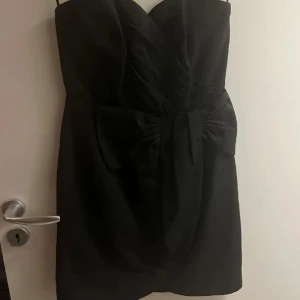 Svart axelbandslös klänning med rosett - Snygg svart axelbandslös klänning med en stor rosett framtill i midjan. Klänningen har en kort längd och en elegant, figurnära passform som framhäver siluetten. Perfekt för dig som vill ha en stilren look med en twist.