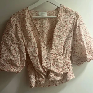 Blommig omlottblus från Neo Noir - Säljer en ljus blommig omlottblus från Neo Noir med puffiga korta ärmar och v-ringad hals. Blusen har små röda och rosa blommor på beige botten, volangdetalj vid midjan och en skön, lite krinklad bomullskänsla. Perfekt för dig som gillar romantisk stil!