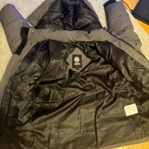 Mörkgrå dunjacka från Canada Goose - Säljer en mörkgrå dunjacka från Canada Goose med huva och klassisk logga på insidan. Jackan har quiltad design, lång ärm och dragkedja framtill. Insidan är fodrad med svart, glansigt material och jackan är perfekt för kalla vinterdagar.