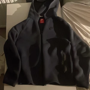 Mörkblå Nike hoodie XL - Snygg mörkblå nike tech hoodie från Nike i storlek XL för barn, med klassisk logga på bröstet och dragkedja framtill. Tröjan har huva och är tillverkad i en mjuk blandning av bomull och polyester. Perfekt för chill eller träning, med långa ärmar och stilren look.