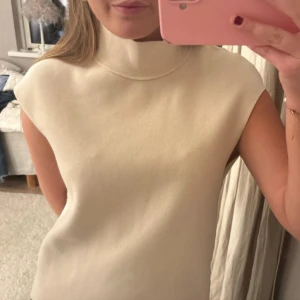 Beige polotopp med hög krage - Säljer en stilren beige polotopp med hög krage och ärmlös design. Toppen har en rak passform och är tillverkad i ett mjukt material som ger en clean och modern look. Perfekt att styla med jeans eller kjol för en enkel men snygg outfit.