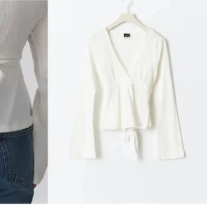 Vit omlottopp med knyt i midjan - Säljer en fräsch vit omlottopp från Gina  med v-ringning och långa ärmar. Toppen har ett snyggt knytband i midjan som ger en feminin siluett och lätt ribbad struktur i tyget. Perfekt att styla med jeans eller kjol för en clean look.