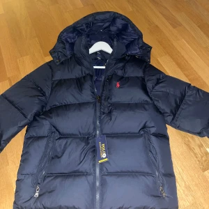 Mörkblå pufferjacka från Polo Ralph Lauren - Snygg mörkblå pufferjacka från Polo Ralph Lauren med huva och dragkedja framtill. Jackan är i storlek M och är inte använd. Har testat den men den var för stor för mig och fick den som en present utan kvitto så kan inte lämna tillbaka.