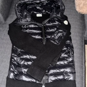 Svart Moncler pufferjacka med huva - Svart pufferjacka från Moncler med stickade ärmar och huva. Jackan har dragkedja framtill, Moncler-logga på ärmen och quiltad kropp i glansigt material. Perfekt för dig som gillar stilren och sportig look.