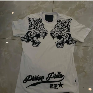 Vit Philipp Plein t-shirt med tigertryck - Snygg vit t-shirt från Philipp Plein med två stora svarta tigerhuvuden tryckta på bröstet och cool logga längst ner. Klassisk rund hals och korta ärmar. Materialet är mjuk bomull som känns skönt mot huden. Perfekt för dig som gillar statement-plagg och streetstyle.