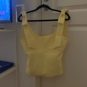 Gul linnetopp med rosetter från Zara - Supersöt ljusgul linnetopp från Zara med breda axelband och fina rosetter på axlarna. Toppen har en fyrkantig halsringning och är figurnära i modellen. Perfekt för sommarens alla tillfällen och ger en fräsch vibe till din outfit.