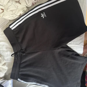 Svarta Adidas shorts med vita ränder - Svarta Adidas shorts med klassiska vita ränder längs sidorna och Adidas-logga framtill. Modellen har snörning i midjan och är tillverkad i mjuk bomull, perfekt för träning eller chill. Snygg och sportig look som alltid funkar.