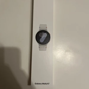 Samsung Galaxy Watch7 Silver -  Helt oöppnad Galaxy Watch7 i silver från Samsung med rund urtavla och stilren design. Smartklockan har en modern look med vit/silverfärgat armband och svart display. Perfekt för dig som vill ha koll på hälsa, träning och notiser direkt på handleden.
