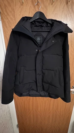 Svart pufferjacka från Canada Goose - Svart pufferjacka från Canada Goose med huva och snygga detaljer som metallspänne och logopatch på ärmen. Jackan har två stora fickor fram, dragkedja och knappar. Perfekt för kalla dagar och har en clean, modern look. (Pris. Kan diskuteras vid snabb affär) 