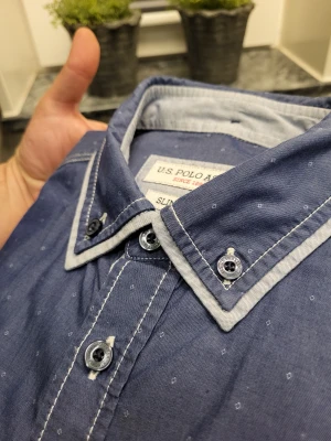 Blå slim fit skjorta från U.S. Polo Assn. - Snygg blå skjorta från U.S. Polo Assn. i slim fit-modell med diskret prickigt mönster. Skjortan har vita kontrastsömmar, knappstängning och detaljer vid krage och bröstficka. Perfekt för dig som gillar stilrena och moderna plagg.