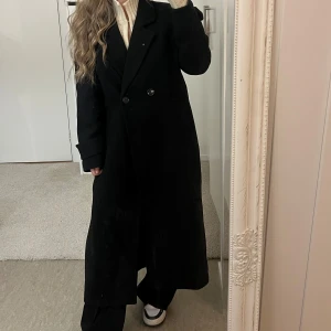 Svart lång kappa med knappar - En lång kappa i vävd kvalitet. Klassisk krage och dubbelknäppning framtill. Kappan har två fickor, långa ärmar med knappdetalj vid armslut. Oversized modell med en rak, loose passform. Perfekt för dig som gillar stilrena och tidlösa plagg. Använd en gång. Slutsåld, nypris 1200 kr.