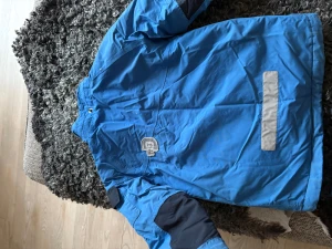 Blå vinterjacka med termobyxor från Didriksons 1913 - Blå vinterjacka och termobyxor från Didriksons 1913 med hög krage, svarta detaljer på axlar och ärmar samt reflexdetaljer. Jackan har dragkedjor på fickorna och justerbara ärmslut. Perfekt för kalla och blåsiga dagar, med slitstarkt material och sportig look. Byxorna matchar jackan och har slitstarka knän och är förstärkt över rumpan med en extra ficka fram.