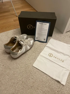 Vita och grå sneakers från Skolyx -  Går ner i pris lätt om ni köper fort, Snygga sneakers från Skolyx i vitt och ljusgrått med klassisk design. Skorna har snörning, rund tå och ovandel i mocka och skinn. Medföljer originalkartong och dustbag med Skolyx-logga. Perfekta för dig som gillar stilrena och tidlösa sneakers.