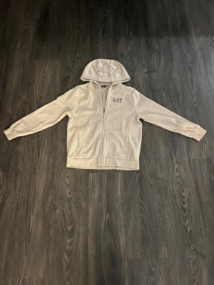 Beige ziphoodie från EA7 Emporio Armani - Snygg beige ziphoodie från EA7 Emporio Armani med huva och dragkedja framtill. Jackan har två fickor och en stilren logga på bröstet. Perfekt för en chill och sportig look. Använd ca 2 gånger.
