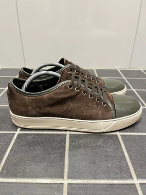 Lanvin - Snygga bruna lanvin sneakers med ovandel i mocka och detaljer i mörkgrönt lack, inklusive tå och snörning. Skorna har rund tå, platt vit sula och mörkgröna kanter. Perfekt för dig som gillar stilrena och unika sneakers med coola materialmixar. Storlek UK6 vilket motsvarar 41/42. Skick 9/10. Endast skorna medföljer