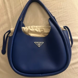 Blå handväska - Snygg blå handväska med rundad form och silverfärgad logga framtill. Väskan har både handtag och avtagbar axelrem, samt en beige innerpåse med snörning. Perfekt för dig som vill ha en stilren och lyxig accessoar. Aldrig använd.