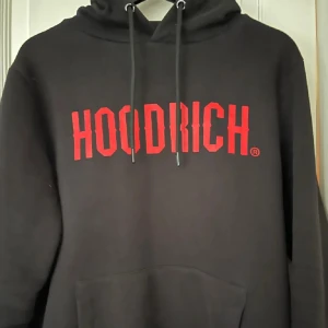 Svart hoodie från Hoodrich med röd logga - Säljer en svart hoodie från Hoodrich med stor röd logotyp på bröstet. Tröjan har huva med dragsko och en stor magficka. Perfekt för en avslappnad och cool stil.