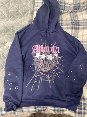 Blå hoodie med spindelnätsprint - Säljer en mörkblå hoodie med trycket 'Atlanta' i rosa och ett stort spindelnätsmotiv med vita stjärnor på bröstet och ärmarna. Tröjan har huva med snörning, magficka och är tillverkad i mjuk fleece. Perfekt för dig som gillar streetwear och coola prints.