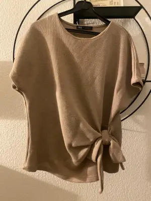 Säljer en beige topp från Zara i storlek S. Toppen har korta ärmar, rund halsringning och en snygg knytdetalj i sidan. Materialet är mjukt och lätt stickat, perfekt för en stilren och enkel look.