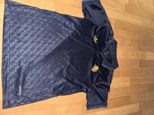 Juventus blå fotbollströja Adidas (XS) - Juventus fotbollströja från Adidas i mörkblått med subtilt geometriskt mönster. Tröjan har korta ärmar, krage med knapp och guldiga detaljer som Adidas-logga, Juventus-text och klubbmärke. Tillverkad i lätt, ventilerande Aeroready-material.