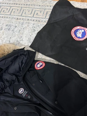 Svart dunjacka Canada Goose med päls - Säljer en svart dunjacka från Canada Goose i storlek M. Jackan har en fluffig huva med naturfärgad päls, quiltad design och klassisk logga på ärmen. Utrustad med dragkedja och knappar framtill. Perfekt för kalla vinterdagar.
