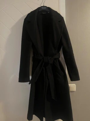 Svart kappa med bälte  - Stilren svart kappa med klassisk krage och knytbälte i midjan. Lång modell med raka ärmar och snygg siluett som ger en clean look.   