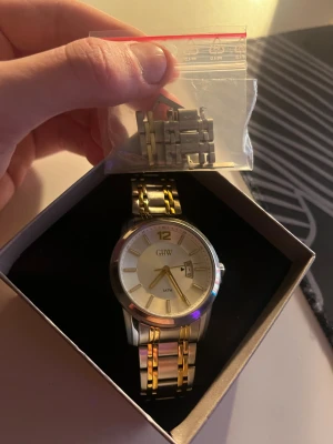 GHW herrklocka i silver och guld - Stilren herrklocka från GHW med rund urtavla i silver och guldtoner. Armbandet är i metall med snygga guldfärgade detaljer och silverlänkar. Klockan har datumvisning och levereras i originalask från Guldfynd samt extra länkar för justering. Finns lite repor på den men inget man ser om man inte kollar noga på den säljes billigt då den e köpt för 1,1k