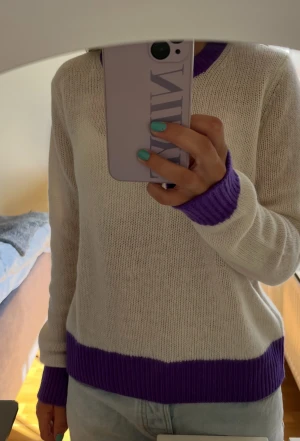 Vit stickad tröja med lila detaljer - Vit stickad tröja från From Future med lila ribbade detaljer vid halsringning, ärmslut och nederkant. Tröjan har en klassisk rund hals och långärmad passform. Perfekt för dig som gillar stilrena färgkombinationer och mjuka material.