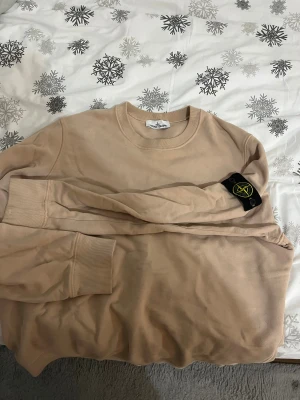 Beige sweatshirt från Stone Island - Säljer en beige långärmad sweatshirt från Stone Island med den klassiska svarta patchen med logga på vänster ärm. Tröjan har rund hals och ribbade muddar vid ärmslut och nederkant. Perfekt för dig som gillar stilrena och exklusiva streetwear-plagg.