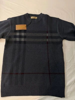 Burberry Sweater - Snygg mörkblå stickad tröja från Burberry med klassiskt rutigt mönster i grått och vinrött på framsidan. Tröjan har rund hals, ribbade muddar och långärmad passform. Perfekt för dig som gillar stilrena designer och ikoniska märken. Storleken passar M, eventuellt L då den är lite lång