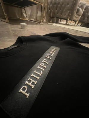 Svart Philipp Plein sweatshirt - Svart sweatshirt från Philipp Plein med metalliskt logotypmärke framtill och ett PP-emblem i guld på ryggen vid nacken. Tröjan har rund hals, ribbade muddar och är tillverkad i mjuk bomull. Snygg och stilren design som sticker ut med exklusiva detaljer.