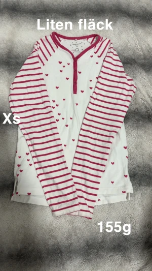 Vit och rosa randig pyjamatröja XS - Söt pyjamaströja från Polarn O. Pyret i vitt med rosa ränder på ärmarna och små hjärtan över hela tröjan. Rund hals med rosa kant och knappar framtill. Perfekt för dig som gillar gulliga detaljer och mjuk bomull.🌸Liten fläck. Storlek xs