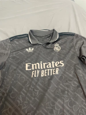 Grå fotbollströja - Snygg grå  fotbollströja från Adidas med diskret mönster, broderat klubbmärke och Adidas-logga på bröstet. Klassisk krage med vit detalj och tre ränder på axlarna. Tillverkad i lätt och ventilerande Aeroready-material.