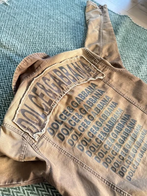Tvärfet Dolce & Gabbana jacka🔥 - Snygg unik beige jeansjacka från Dolce & Gabbana med tryckt logga i flera rader på ryggen och patchade logodetaljer fram. Klassisk krage, två bröstfickor och knäppning med silvriga knappar. Jackan har en cool, streetig vibe och är tillverkad i bomull. Oerhört niche och rare!