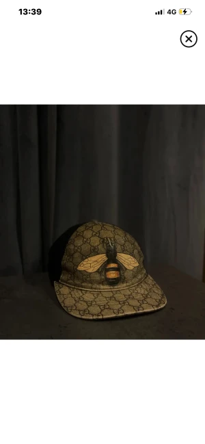 Gucci keps  - Snygg keps från Gucci med klassiskt GG-mönster i beige och brunt framtill, svart mesh på sidor och bak samt broderad bi på framsidan. Justerbar rem med grönt och rött band bak. Märkt med Gucci på insidan. Perfekt för dig som vill sticka ut.