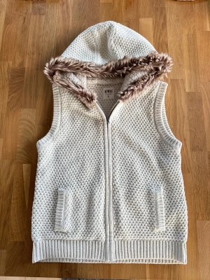 Vintage stickad väst med päls🐻 - Mysig vit stickad väst med huva och päls runt kanten. Västen har grovstickad struktur, två fickor framtill och stängs med dragkedja. Insidan av huvan är fodrad med mjukt teddytyg för extra komfort. Perfekt för lager-på-lager under kyliga dagar. Dam storlek xl herr storlek s/m