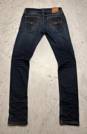 Mörkblå Nudie Jeans - Snygga mörkblå jeans från Nudie Jeans med klassisk orange söm på bakfickorna. Jeansen har en snygg fade som skapats med tiden. Jeansen är storlek W29 L34 och modellen ”tight long John” men jag skulle säga att de passar mer som W29 L32. Skriv om du har frågor!