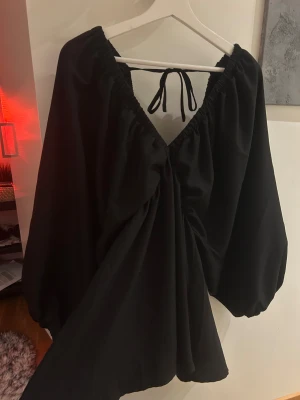 Svart omlottklänning med knyt - Snygg svart omlottklänning med v-ringad hals och knyt i ryggen. Klänningen har långa puffärmar och ett flowigt fall som ger en chill vibe. Perfekt för dig som gillar stilrena plagg med coola detaljer. Materialet känns lätt och bekvämt mot huden.