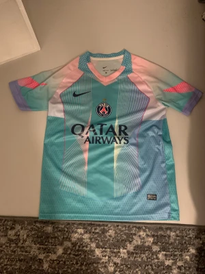 PSG  fotbollströja - Säljer en Paris Saint-Germain fotbollströja från Nike i snygga pastellfärger. Tröjan har korta ärmar, v-ringning och coola grafiska linjer i turkos, rosa och lila. Materialet är lätt och andas, perfekt för fotboll eller streetstyle. PSG-logga och Qatar Airways-tryck på bröstet.