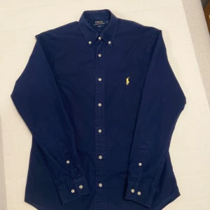Mörkblå skjorta från Polo Ralph Lauren - Snygg mörkblå skjorta från Polo Ralph Lauren med klassisk button-down krage och vita knappar. Ikoniska broderade loggan i gult på bröstet. Skjortan har lång ärm och är tillverkad i mjuk bomull som känns skön mot huden.