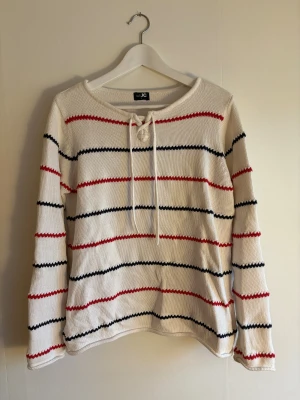 Randig stickad tröja från JC - Vit stickad tröja från JC med horisontella ränder i rött och svart. Tröjan har lång ärm och snörning vid halsen för en avslappnad look. Tillverkad i 100% bomull vilket gör den mjuk och skön att bära.