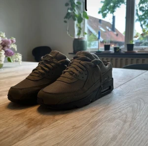 Nike Air Max 90 olivgröna sneakers - Säljer ett par Nike Air Max 90 sneakers i olivgrönt med svart sula. Skorna har en klassisk siluett med rund tå, snörning och meshdetaljer för extra ventilation. Perfekt för dig som gillar streetwear och vill ha en snygg och bekväm sneaker med ikonisk Air-dämpning.