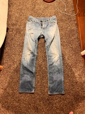 Tommy Hilfiger Denim Ryder jeans W34 L34 - Snygga ljusblå jeans från Tommy Hilfiger Denim, modell Ryder. De har raka ben, klassisk femficksdesign och slitna detaljer på knä och fickor för en cool vintagekänsla. Jeansen är tillverkade i bomull och har knappgylf.