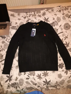 Svart kabelstickad tröja Polo Ralph Lauren - Svart kabelstickad tröja från Polo Ralph Lauren med klassisk röd logobrodering på bröstet. Tröjan har rund hals och långa ärmar, tillverkad i mjuk bomull. Perfekt för dig som gillar stilrena och tidlösa plagg med premiumkänsla.